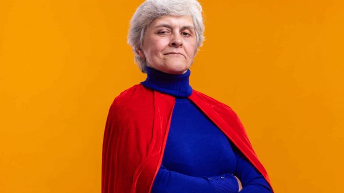 senior woman superhero wearing red cape looking at camera with confident expression with arms crossed standing over orange background