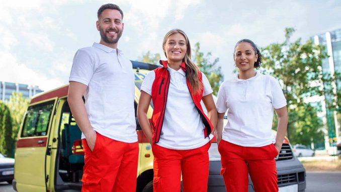 Three multiracial paramedics standing in front of ambulance vehicle, carrying portable equipment